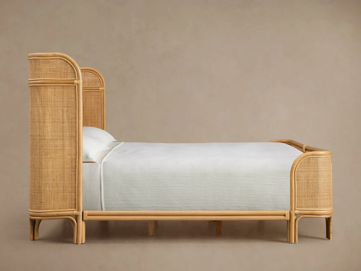 Close-up of Maya Bed headboard, showing light natural rattan with a curved frame and woven mesh panel. Soft, warm tone.
