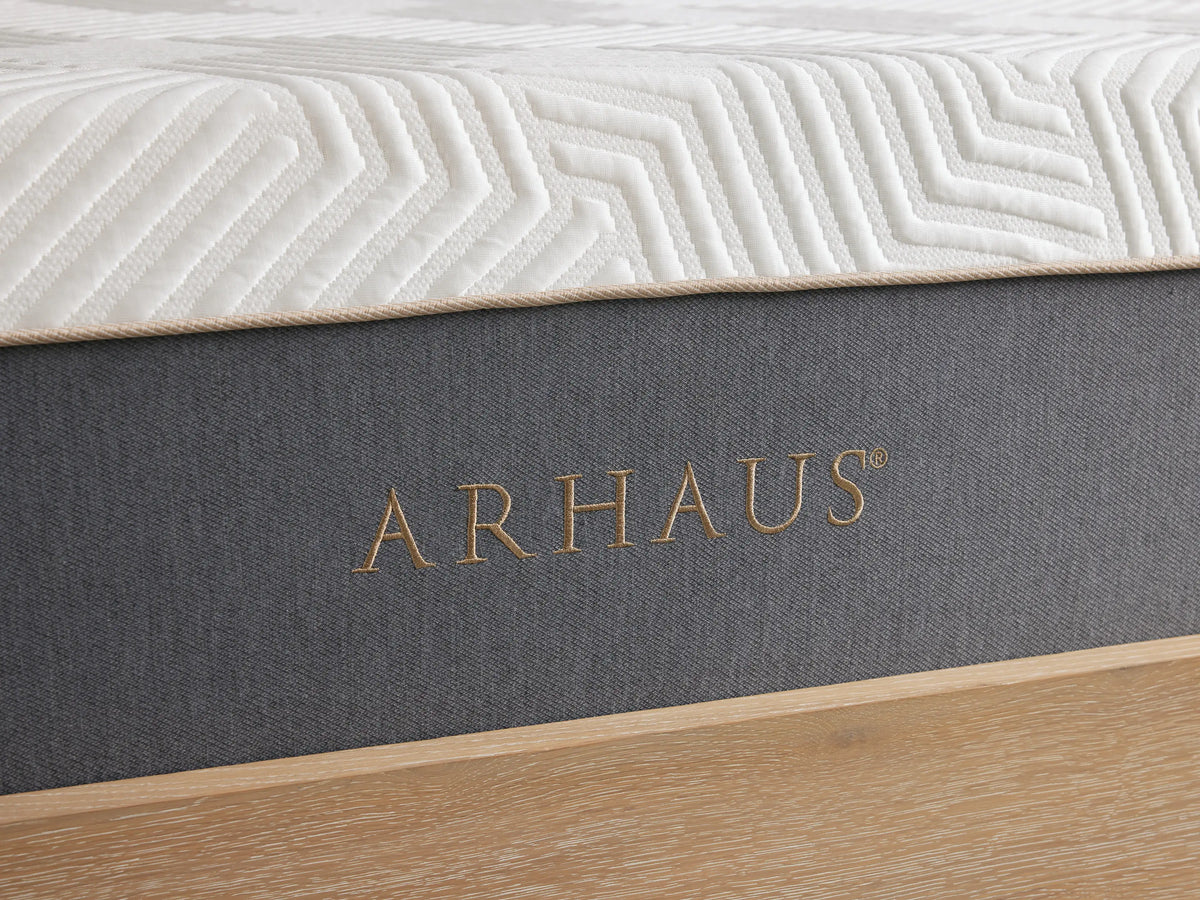 A hand presses down on a white plush mattress with a geometric pattern, showing its softness. The hand wears a delicate bracelet and a ring.