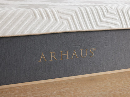 A hand presses down on a white plush mattress with a geometric pattern, showing its softness. The hand wears a delicate bracelet and a ring.