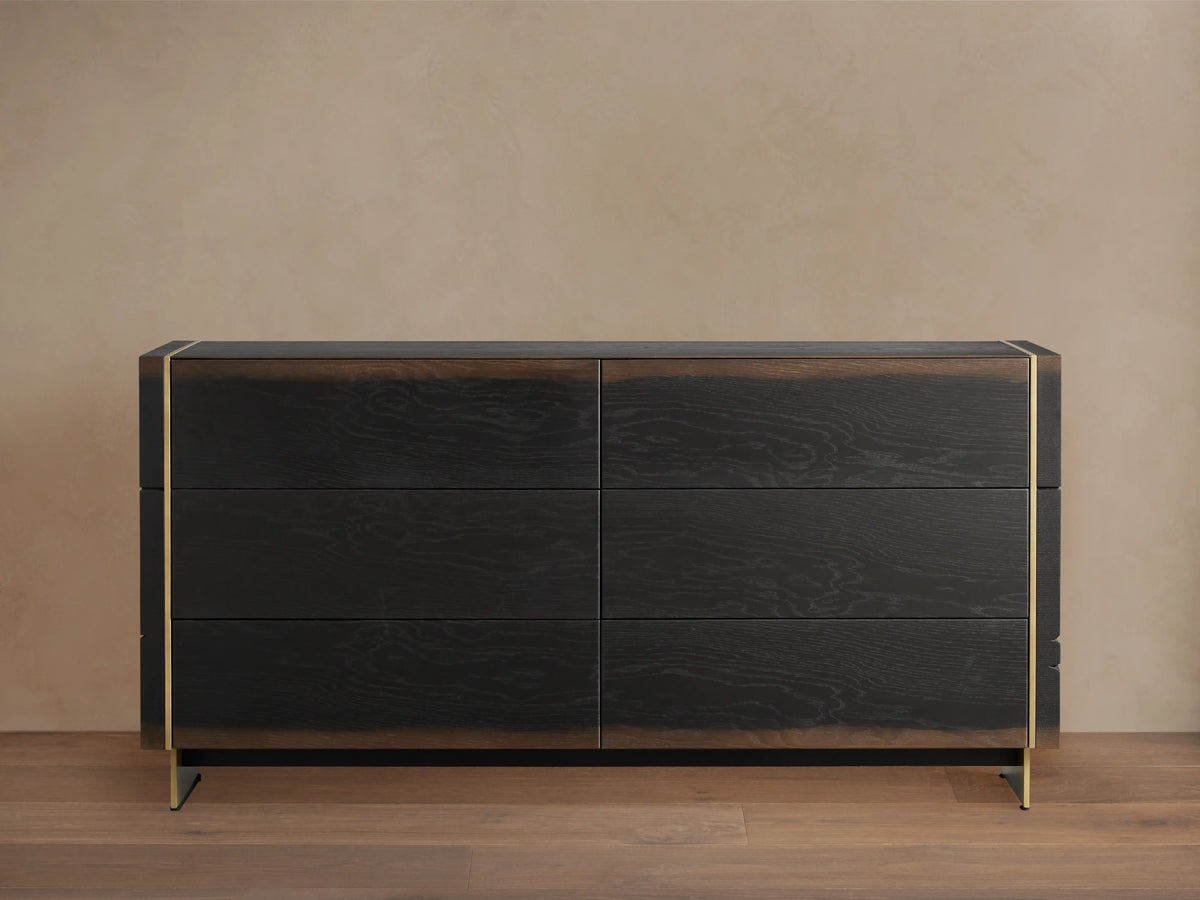 Mihaela Six-Drawer Dresser in dark oak with six drawers, featuring metallic brass accents on the edges, against a beige wall.