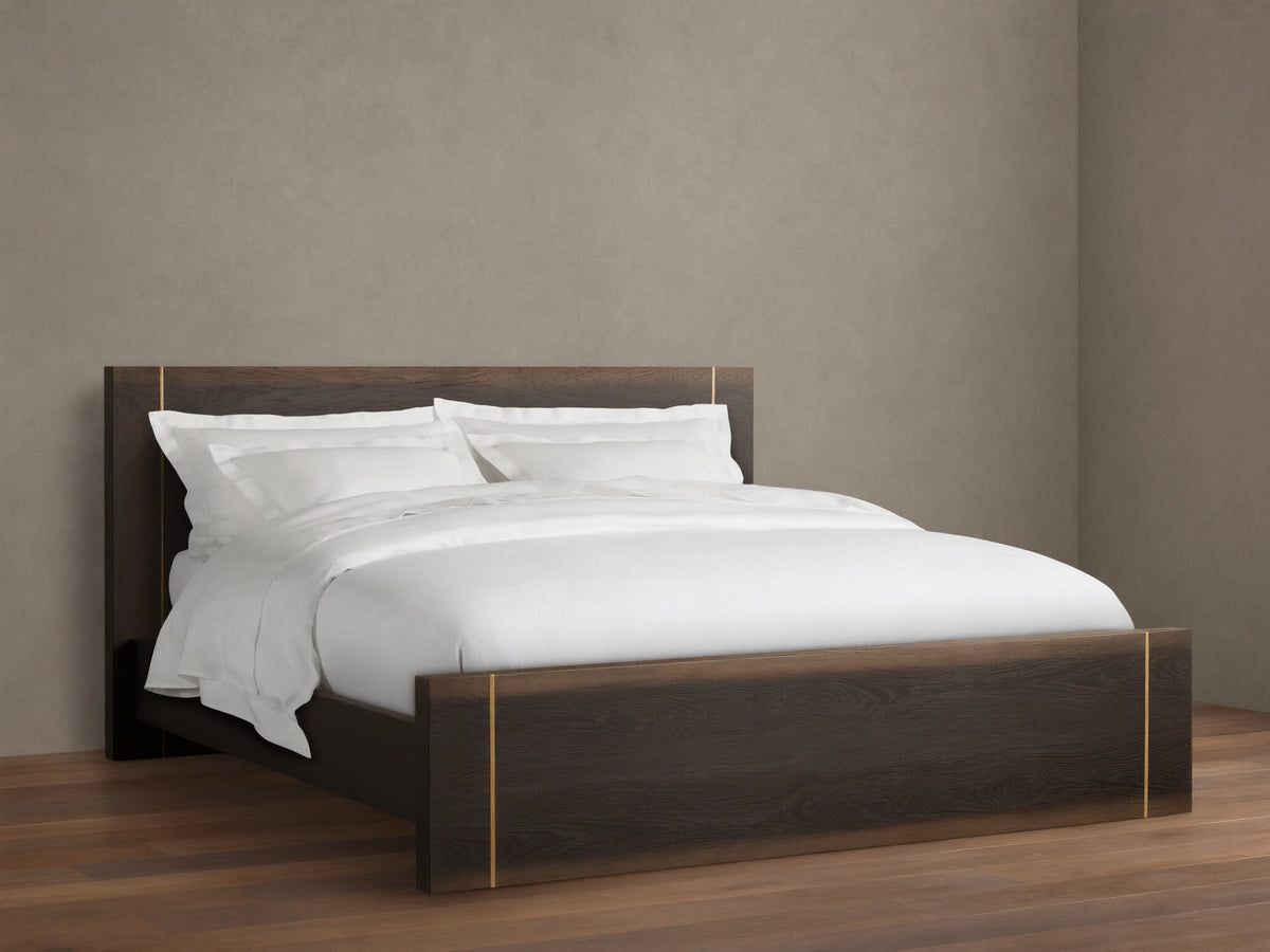 Mihaela Bed with dark oak frame, metallic brass accents, white bedding, on a wooden floor, set against a beige wall.