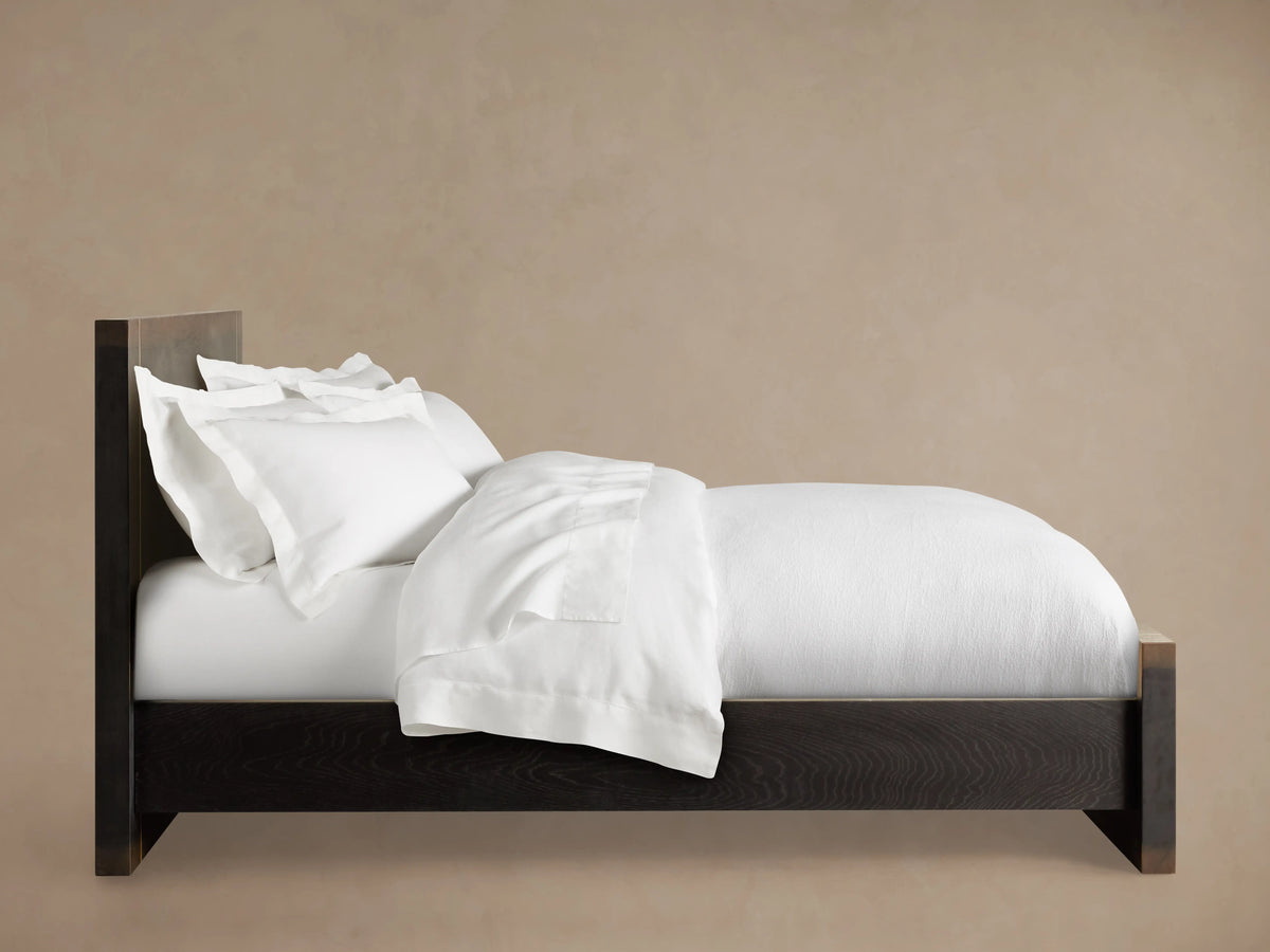 Mihaela Bed with dark oak frame, metallic brass accents, white bedding, on a wooden floor, set against a beige wall.