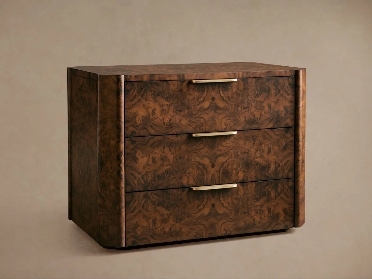 A wooden chest of drawers with three compartments, featuring a dark burl wood finish and sleek metallic handles, placed against a beige wall.
