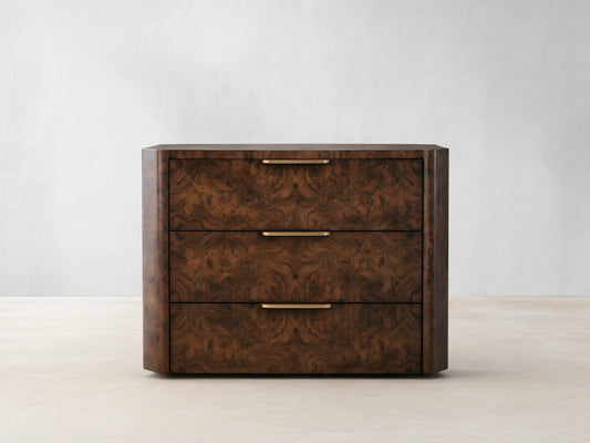 A wooden chest of drawers with three compartments, featuring a dark burl wood finish and sleek metallic handles, placed against a beige wall.