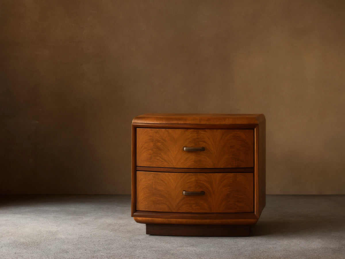Warm brown Palma Nightstand with distinctive cerajeira wood veneer, two drawers, aged bronze hardware, beside a bed.