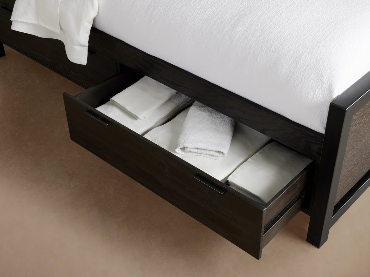 Dark wood Palmer Storage Bed with an open drawer, showing neatly folded white linens inside. Part of the bed frame and bedding are visible.