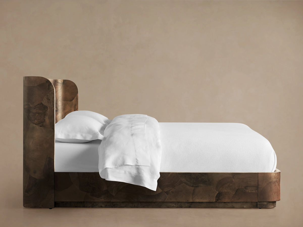 Bed made from dark wood with natural grain pattern, featuring a headboard. White bedding on a sturdy, low frame on wooden floor.