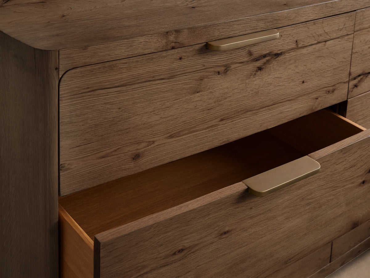 Close-up of a brown oak Redding Six-Drawer Dresser with a lower drawer partially open and gold pull handles.