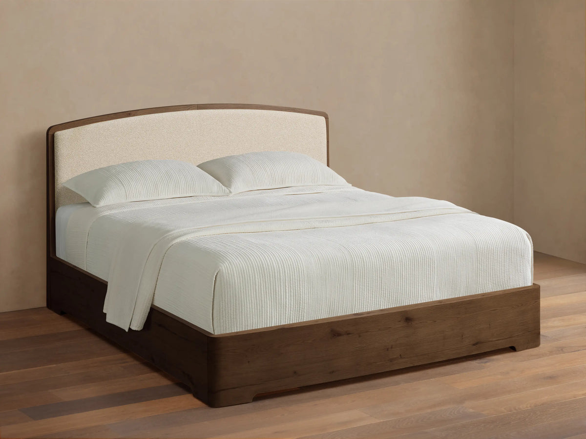 Angled view of the Redding Bed with a dark brown wood frame, cream upholstered headboard, and white vertically ribbed bedding.