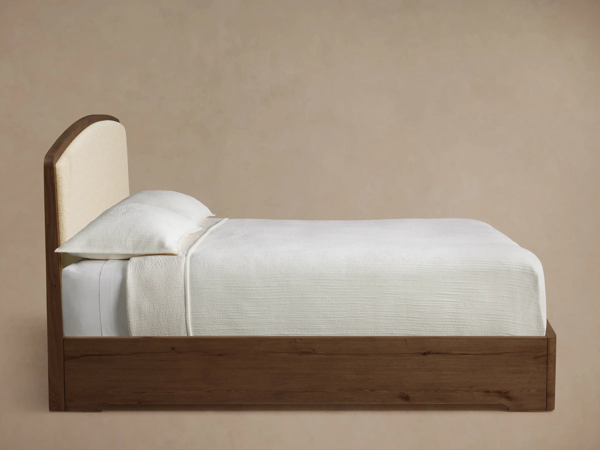 Side profile of the Redding Bed with a dark wood frame and arched cream upholstered headboard, dressed in white bedding.