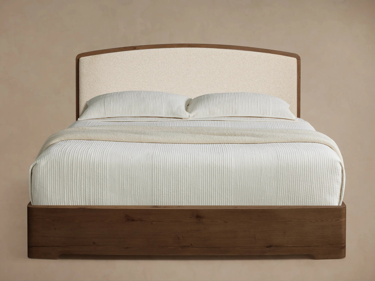 Angled view of the Redding Bed with a dark brown wood frame, cream upholstered headboard, and white vertically ribbed bedding.