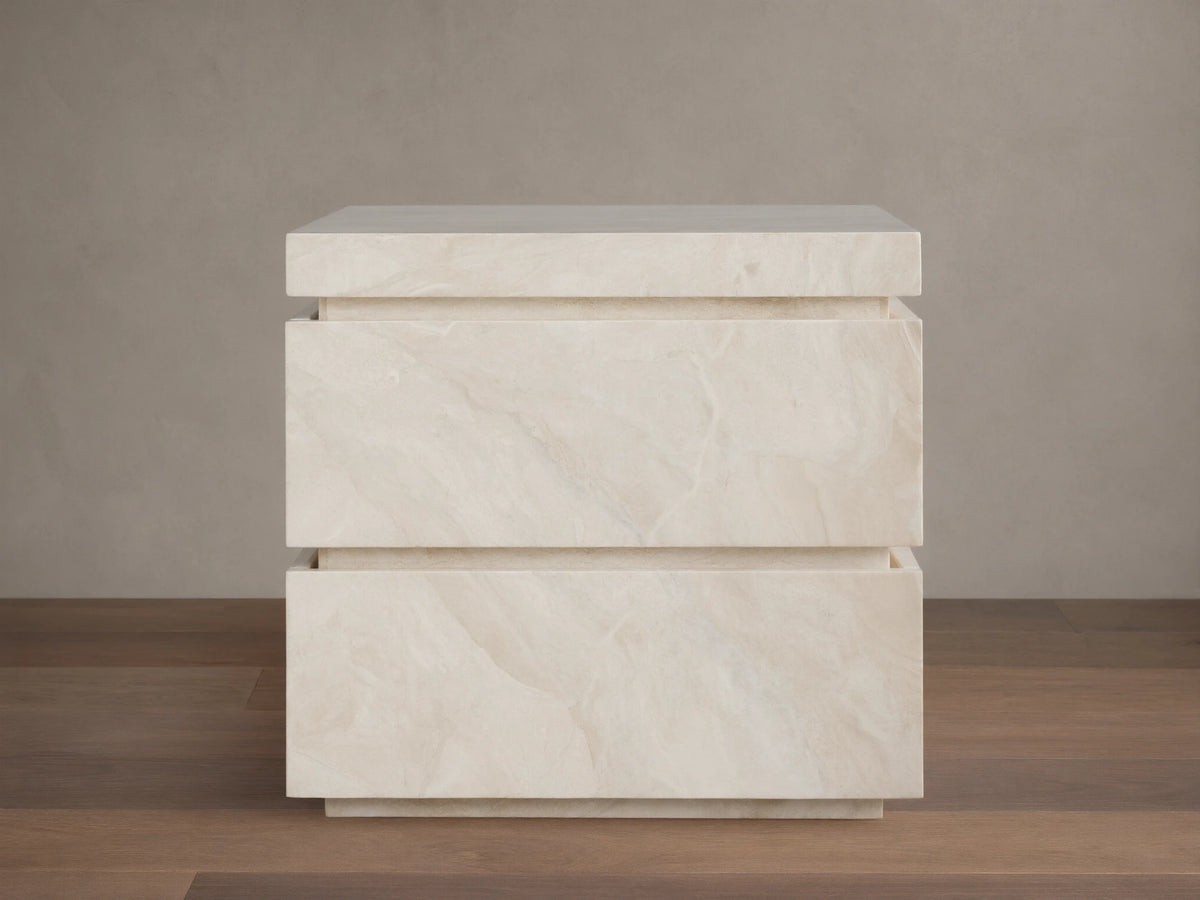 Rive Stone Nightstand, two-drawer, light beige Victoria Falls stone with natural veining, against a beige wall & wooden floor.
