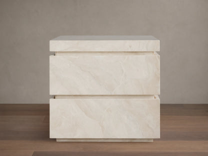 Rive Stone Nightstand, two-drawer, light beige Victoria Falls stone with natural veining, against a beige wall & wooden floor.