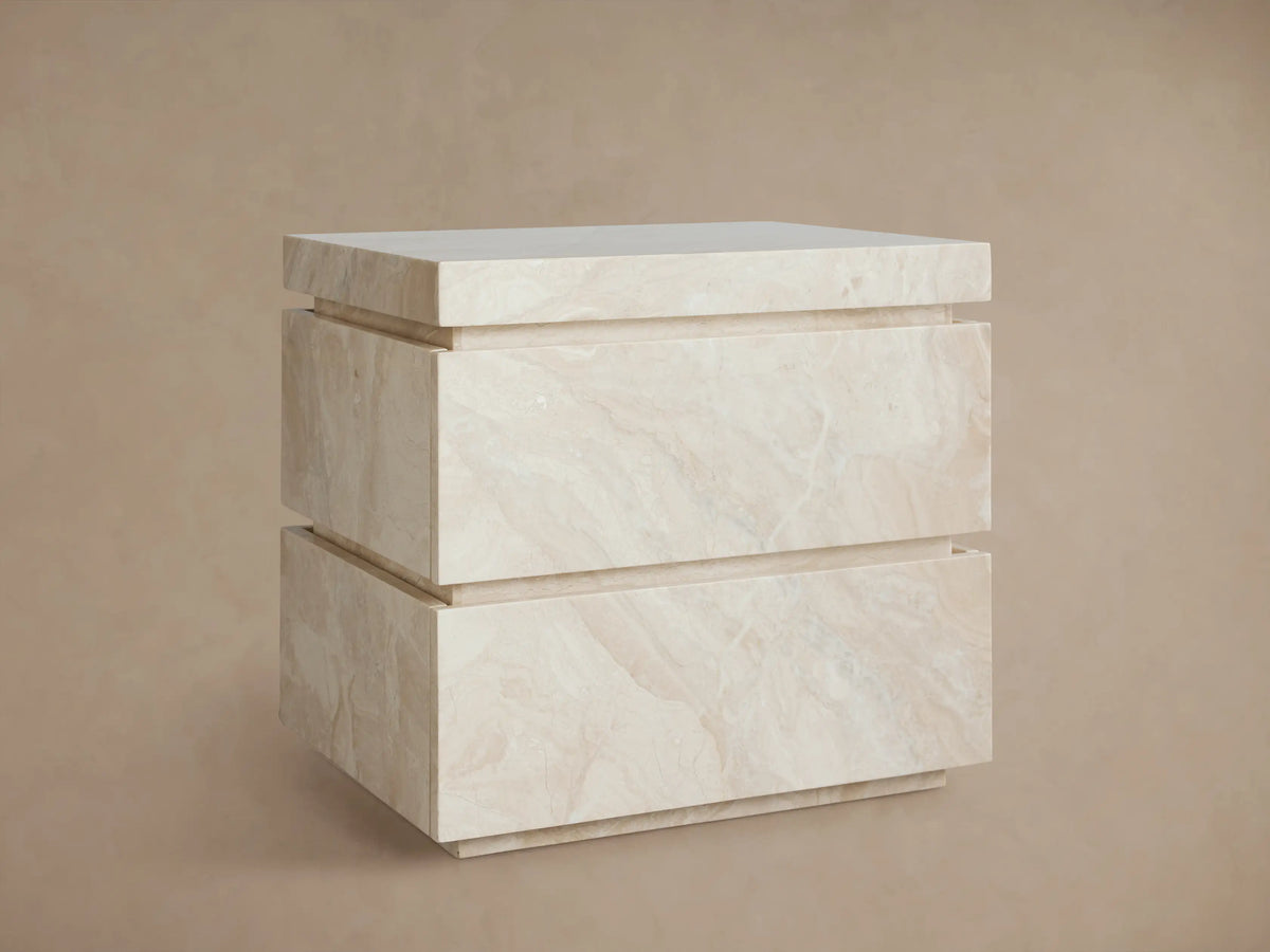Rive Stone Nightstand, two-drawer, light beige Victoria Falls stone with natural veining, against a beige wall & wooden floor.