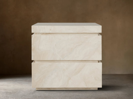 Rive Stone Nightstand, two-drawer, light beige Victoria Falls stone with natural veining, against a beige wall & wooden floor.