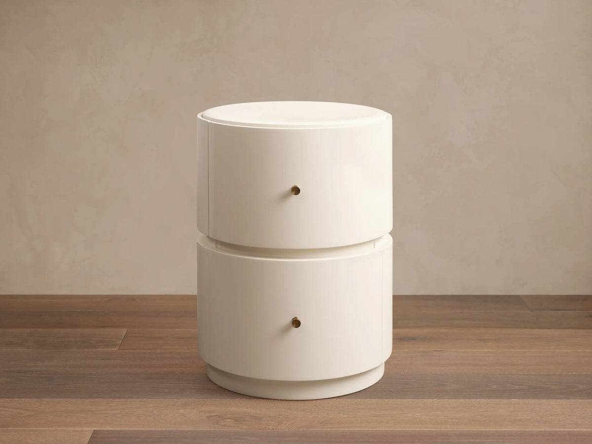 Riya Round Nightstand, off-white, high-gloss, with two drawers & brass knobs, on a wooden floor against a beige wall.