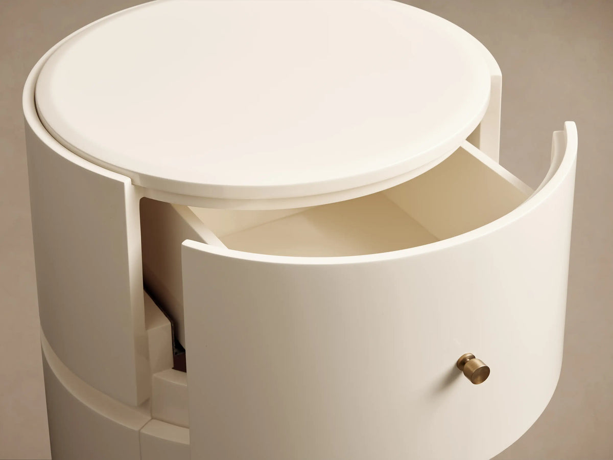 Riya Round Nightstand, off-white, high-gloss, with two drawers & brass knobs, on a wooden floor against a beige wall.