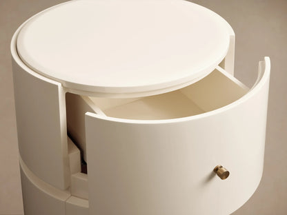Riya Round Nightstand, off-white, high-gloss, with two drawers & brass knobs, on a wooden floor against a beige wall.