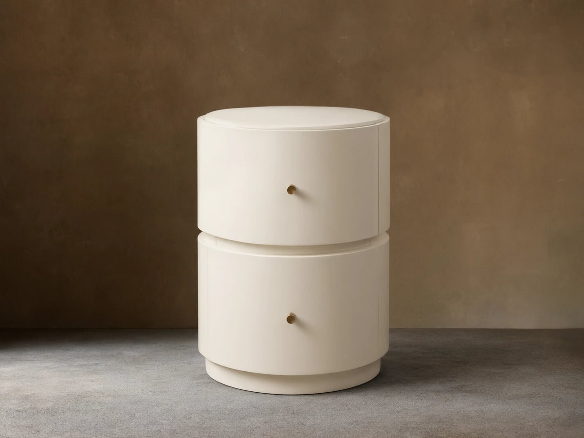 Riya Round Nightstand, off-white, high-gloss, with two drawers & brass knobs, on a wooden floor against a beige wall.