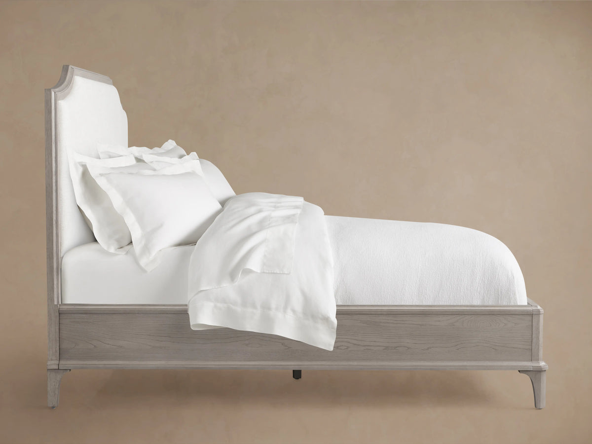 Front view of the St. Martin Bed, featuring an oak wood frame with a light gray finish and a curved, off-white upholstered headboard.