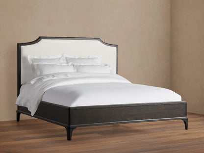 St. Martin Bed with dark oak frame, white upholstered headboard, white bedding, on hardwood floor against beige wall.