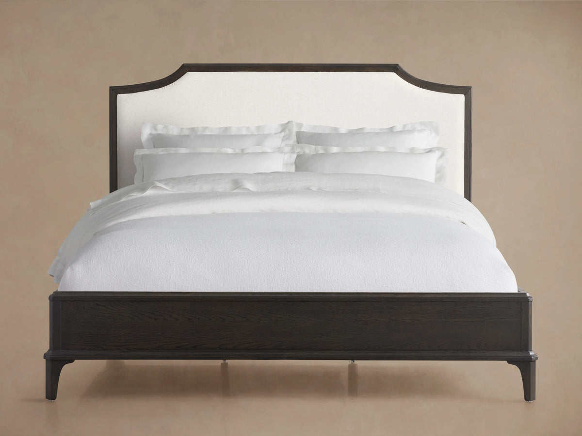 St. Martin Bed with dark oak frame, white upholstered headboard, white bedding, on hardwood floor against beige wall.