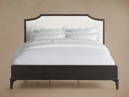 St. Martin Bed with dark oak frame, white upholstered headboard, white bedding, on hardwood floor against beige wall.