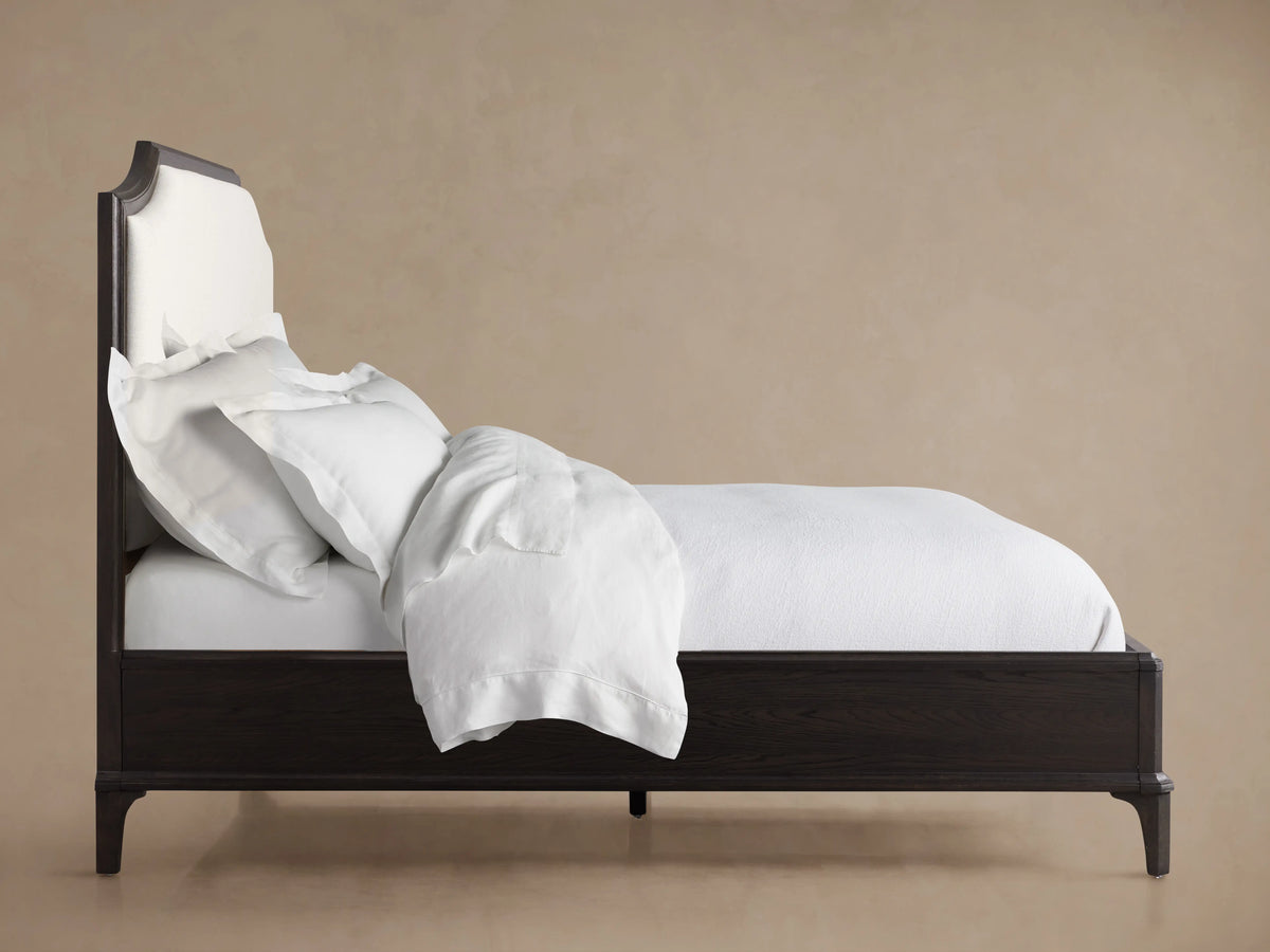 St. Martin Bed headboard with dark oak wood frame and light fabric upholstery, featuring a curved top design against a neutral wall.