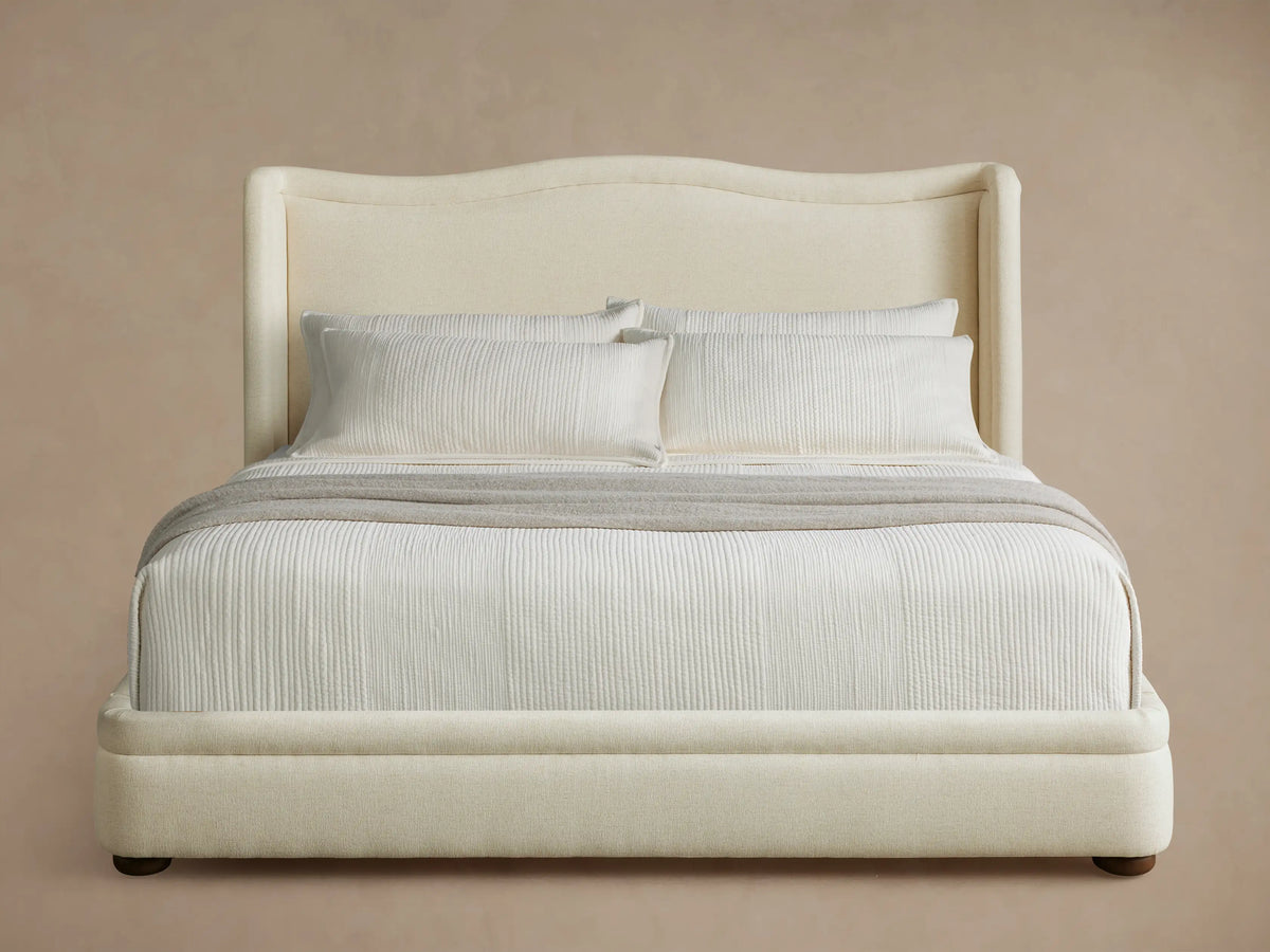 Front view of the Winslet Bed, featuring a tall, curved brown upholstered headboard, dressed with cream bedding and a patterned bench.