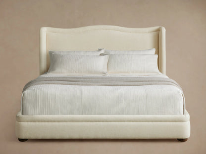 Front view of the Winslet Bed, featuring a tall, curved brown upholstered headboard, dressed with cream bedding and a patterned bench.
