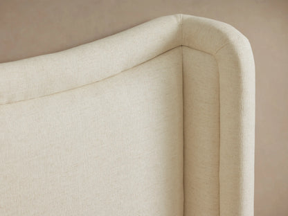 Winslet Bed, a cream upholstered bed with a gently curved, wingback headboard, white ribbed bedding, and a gray throw.