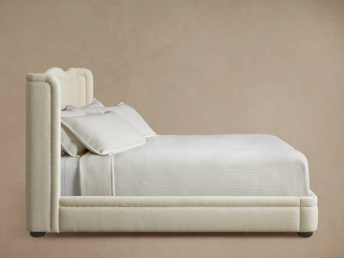 Close-up of the Winslet Bed headboard's top right corner, upholstered in textured, light cream fabric with curved and winged details.