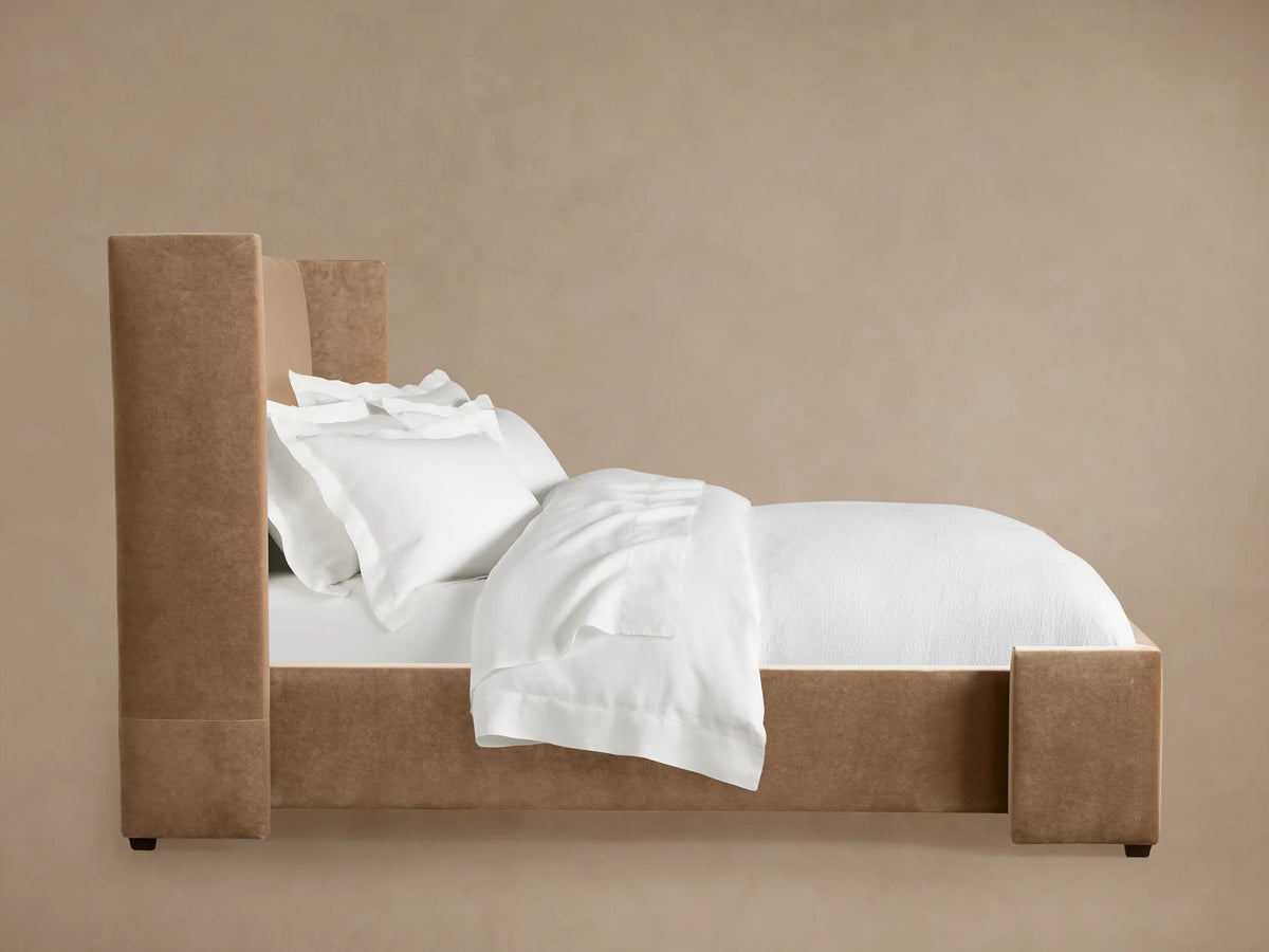 Camel upholstered Wyller Bed with a tall headboard, made up with crisp white sheets, pillows, and duvet against a neutral wall.