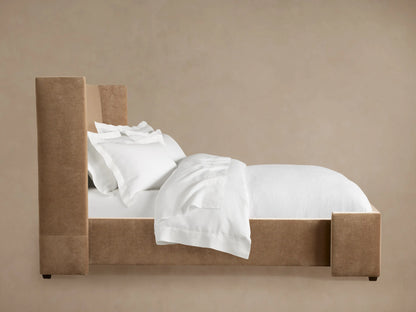 Camel upholstered Wyller Bed with a tall headboard, made up with crisp white sheets, pillows, and duvet against a neutral wall.