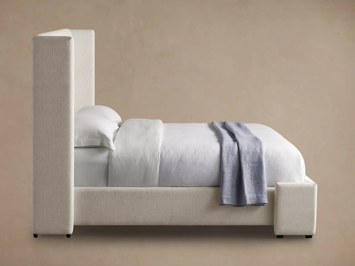 Close-up of Wyller Bed footboard and side rail in light beige textured fabric with white sheets & small wooden leg.