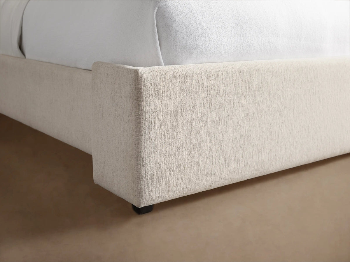 Wyller Bed, upholstered in light beige fabric, with a tall winged headboard. Made up with white bedding and a blue throw.