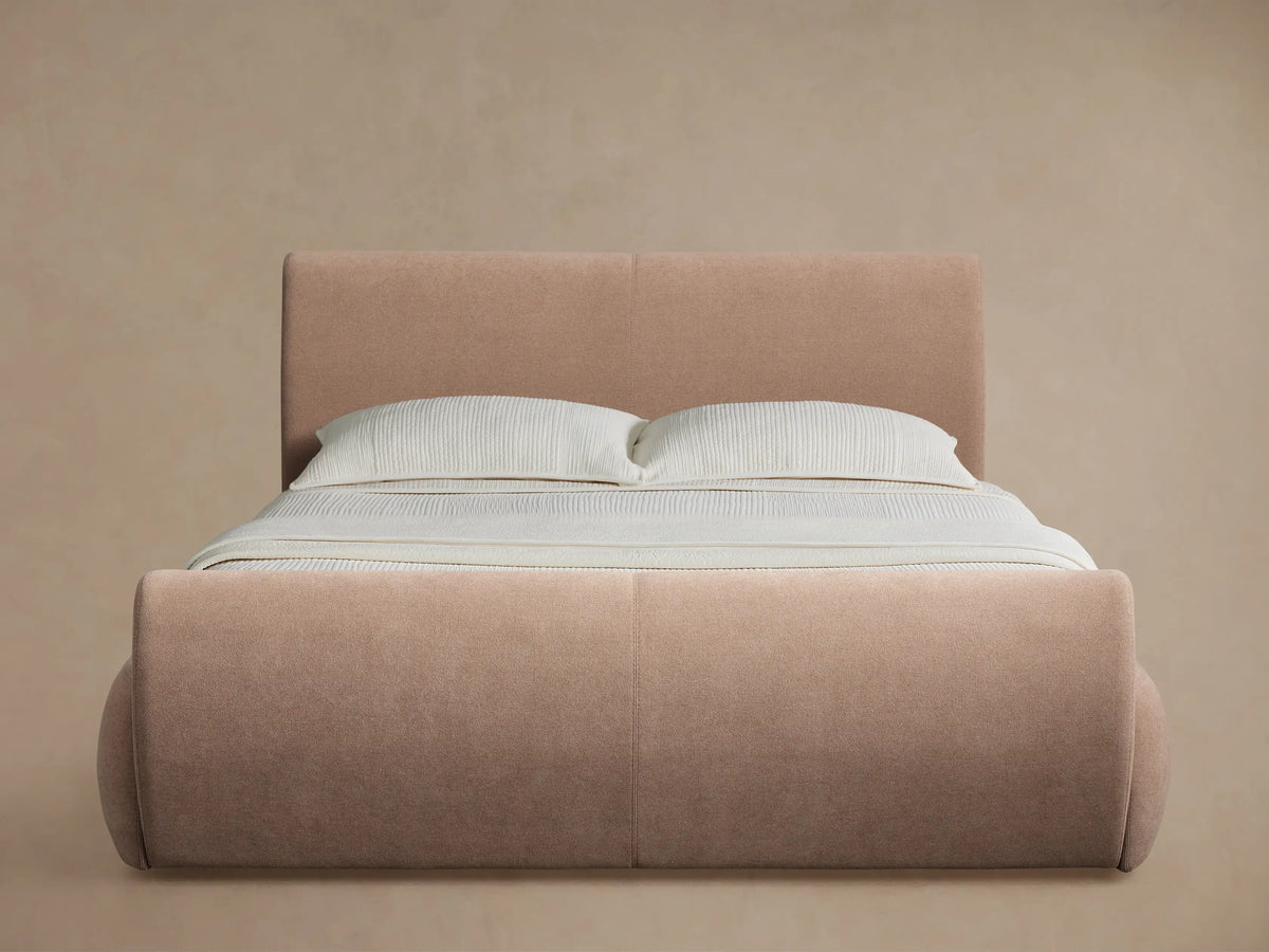 Full view of the Dusty Rose upholstered Wynn Bed, with a curved footboard and white bedding, in a bright bedroom.