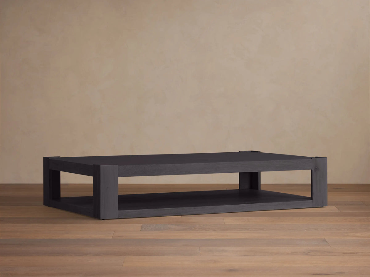 Devon Coffee Table: Dark gray/black rectangular oak table with two shelves, on a wood floor against a beige wall.