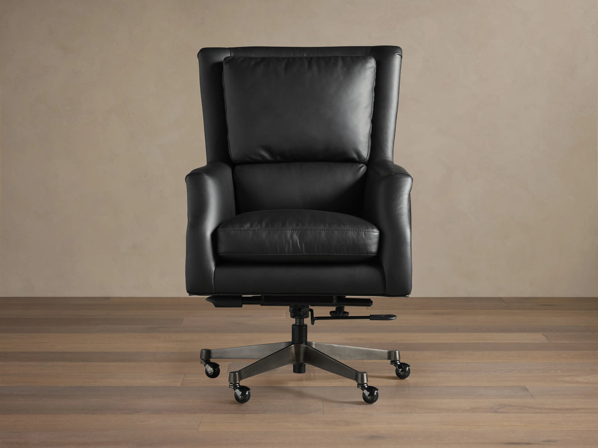 Black leather desk chair on a wooden floor, featuring cushioned seat and back, adjustable base with wheels, and nailhead trim.