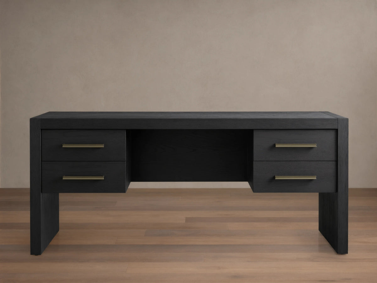 Black Bodhi Executive Desk with visible wood grain, four drawers with brass pulls, centered on a wood floor against a neutral wall.