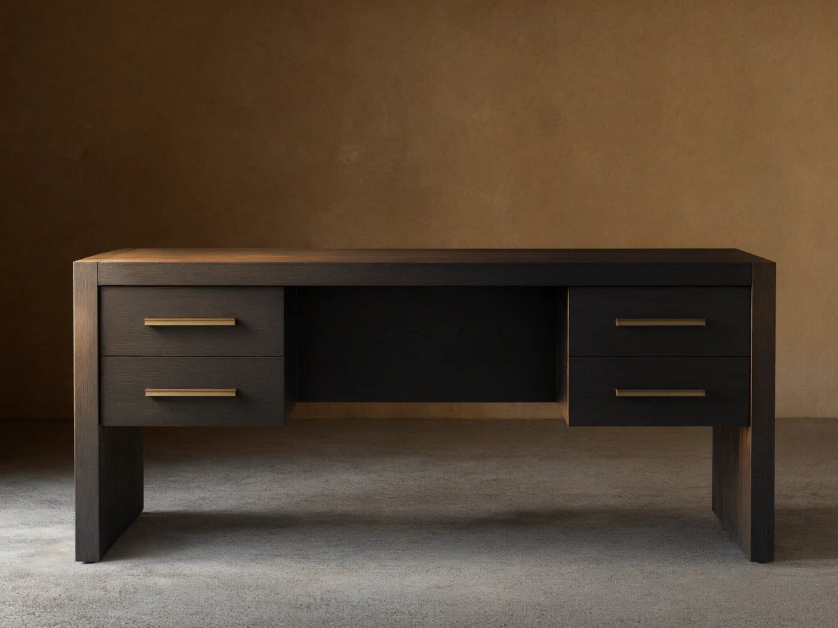 Black Bodhi Executive Desk with visible wood grain, four drawers with brass pulls, centered on a wood floor against a neutral wall.