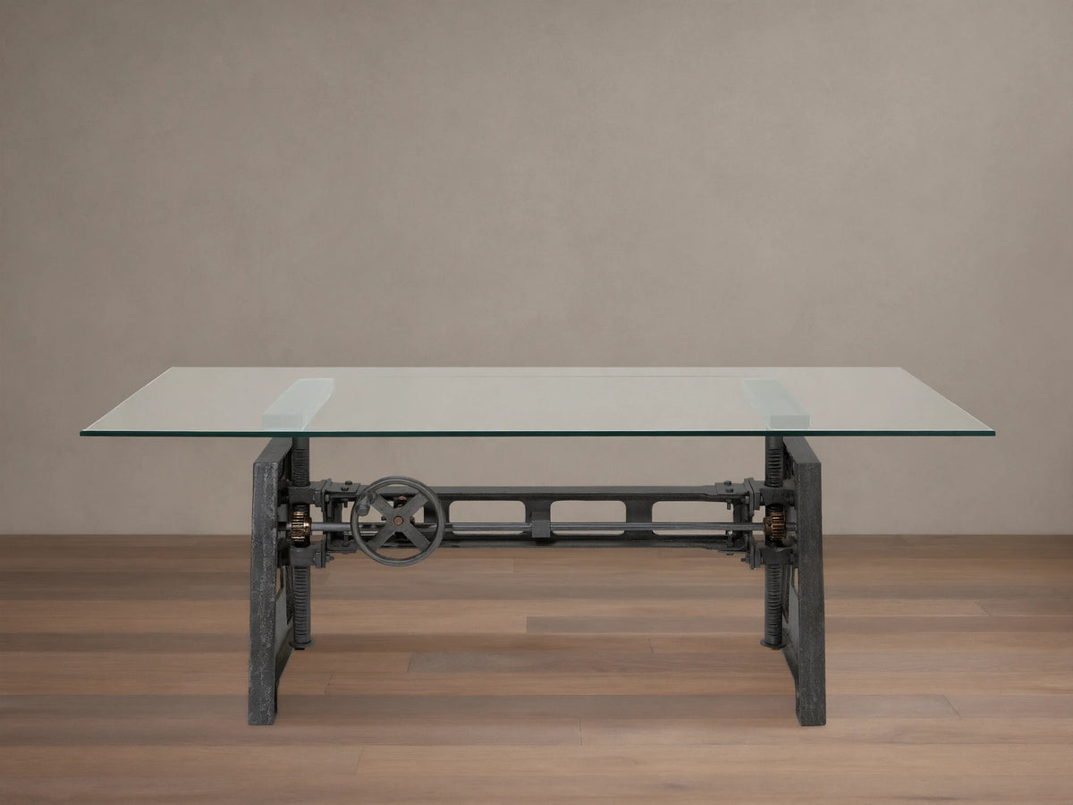 Industrial Crank Desk with clear rectangular glass top, dark metal base featuring a prominent crank wheel, on a wooden floor.