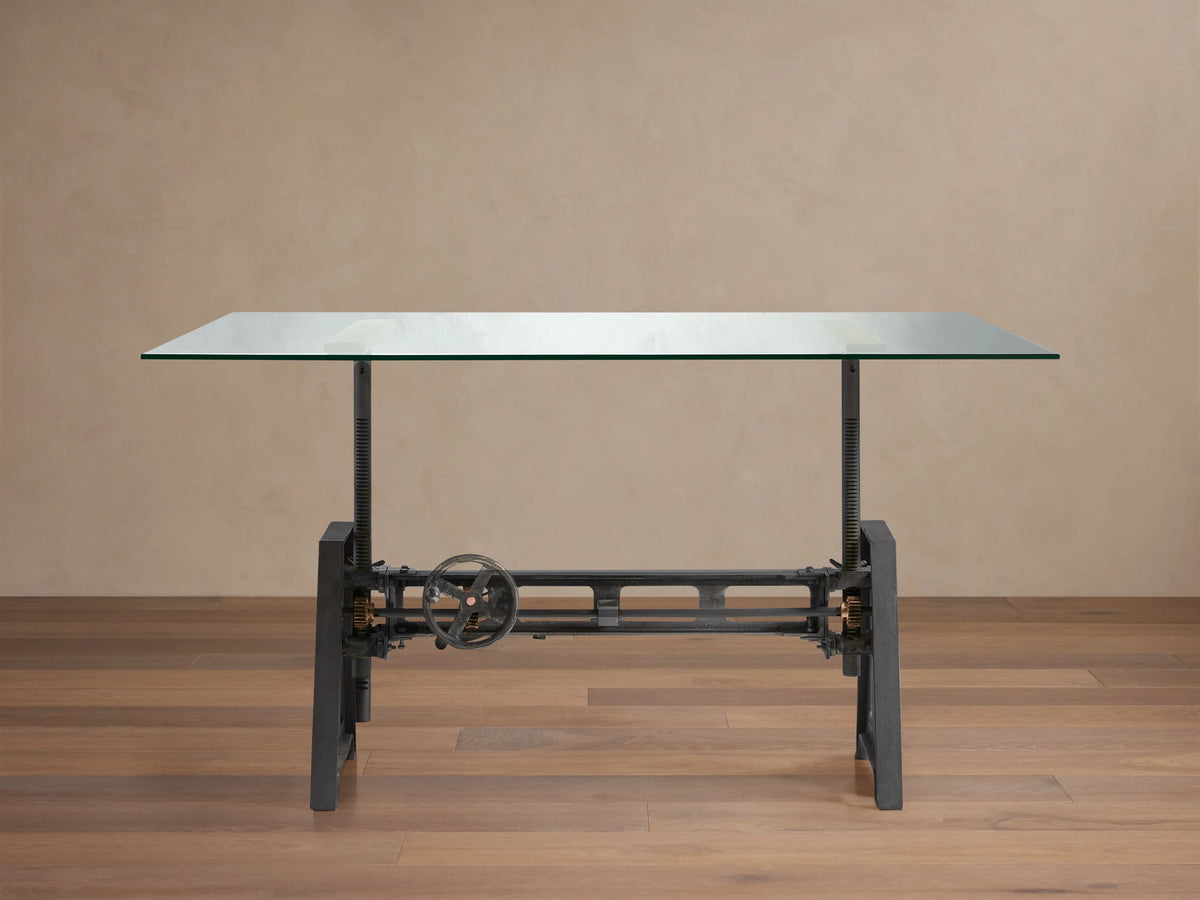 Industrial Crank Desk with clear rectangular glass top, dark metal base featuring a prominent crank wheel, on a wooden floor.