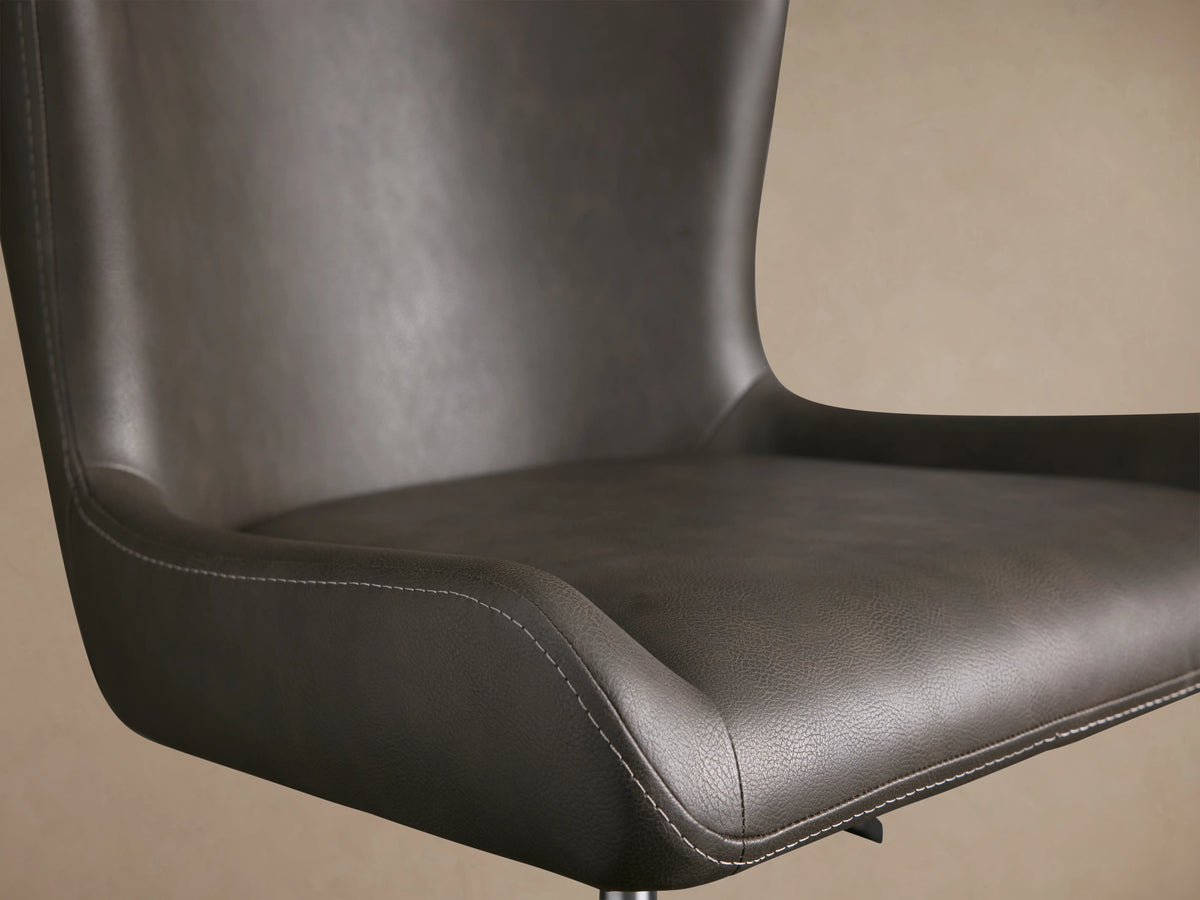 Kirsten Faux Leather Desk Chair with dark upholstery, curved backrest, and visible stitching on seat. Brown background.
