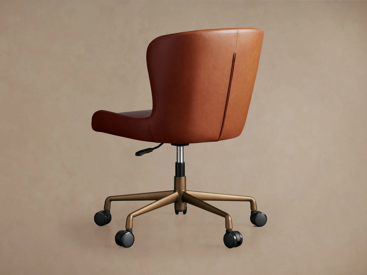 Kirsten Faux Leather Desk Chair, seen from the back, features a warm brown faux leather upholstery, swivel base, and caster wheels on metal legs.