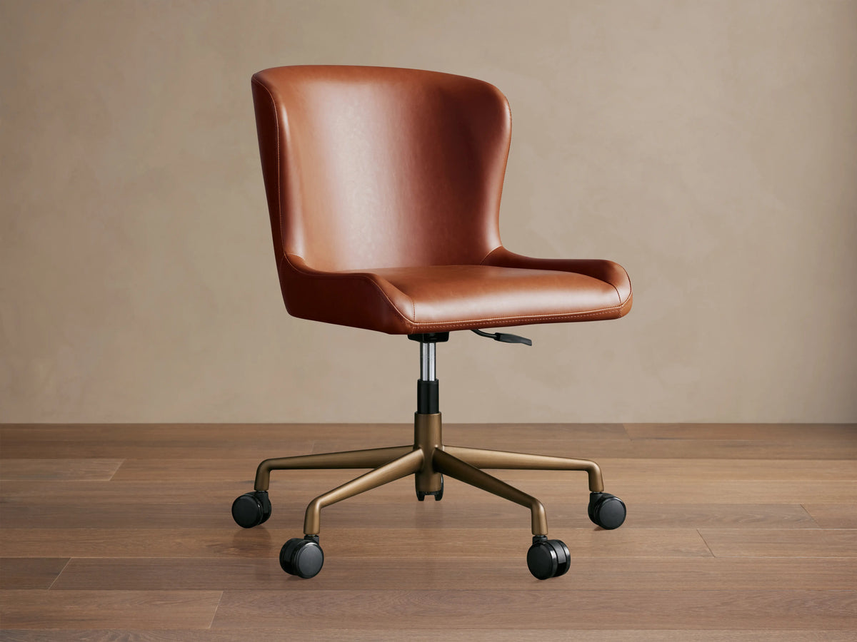 Kirsten Faux Leather Desk Chair in warm brown, features a smooth seat and backrest, gold-toned metal base with black casters.