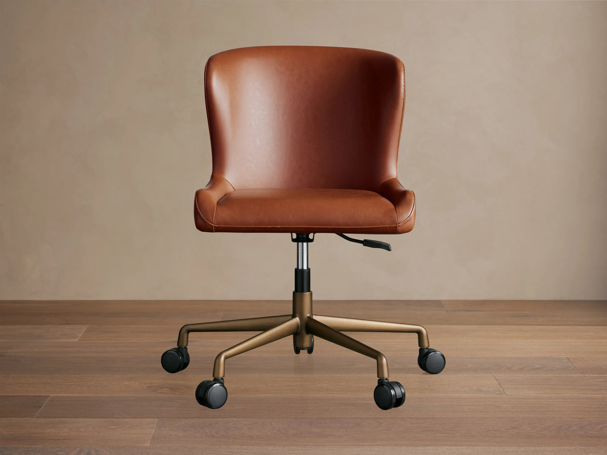 Brown Kirsten Faux Leather Desk Chair with a high back, on a bronze swivel base with casters, set against a wooden floor.