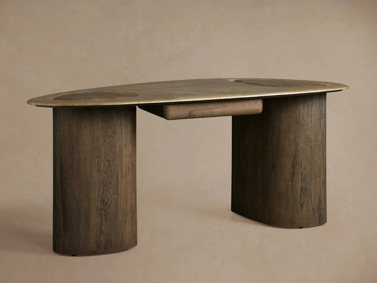 Sucurro Writing Desk with oval oxidized aluminum top & two thick dark wood cylindrical legs, front view, against a beige wall.