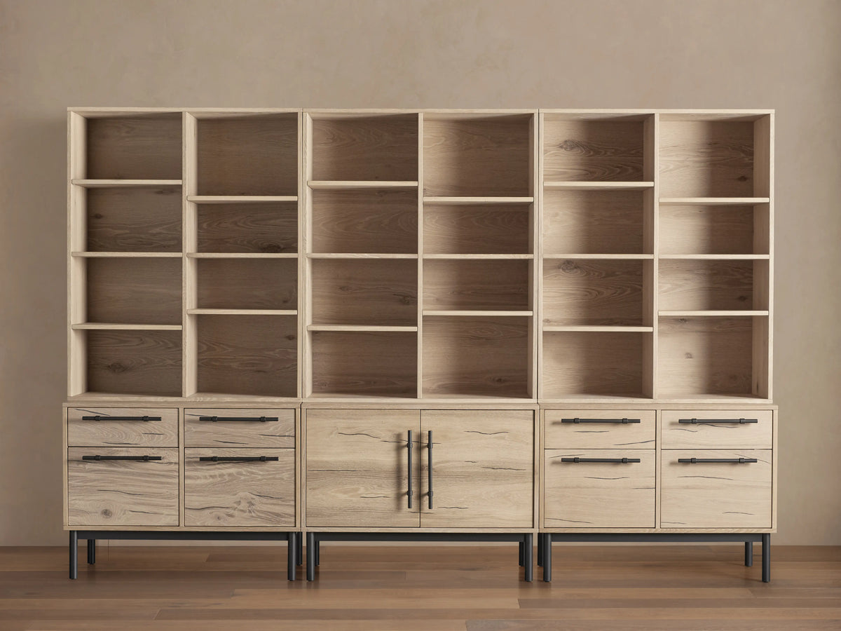 Sullivan Triple Bookcase in natural oak finish, showcasing adjustable shelves and iron-handled drawers. Placed on a wooden floor.