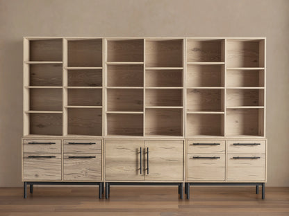 Sullivan Triple Bookcase in natural oak finish, showcasing adjustable shelves and iron-handled drawers. Placed on a wooden floor.